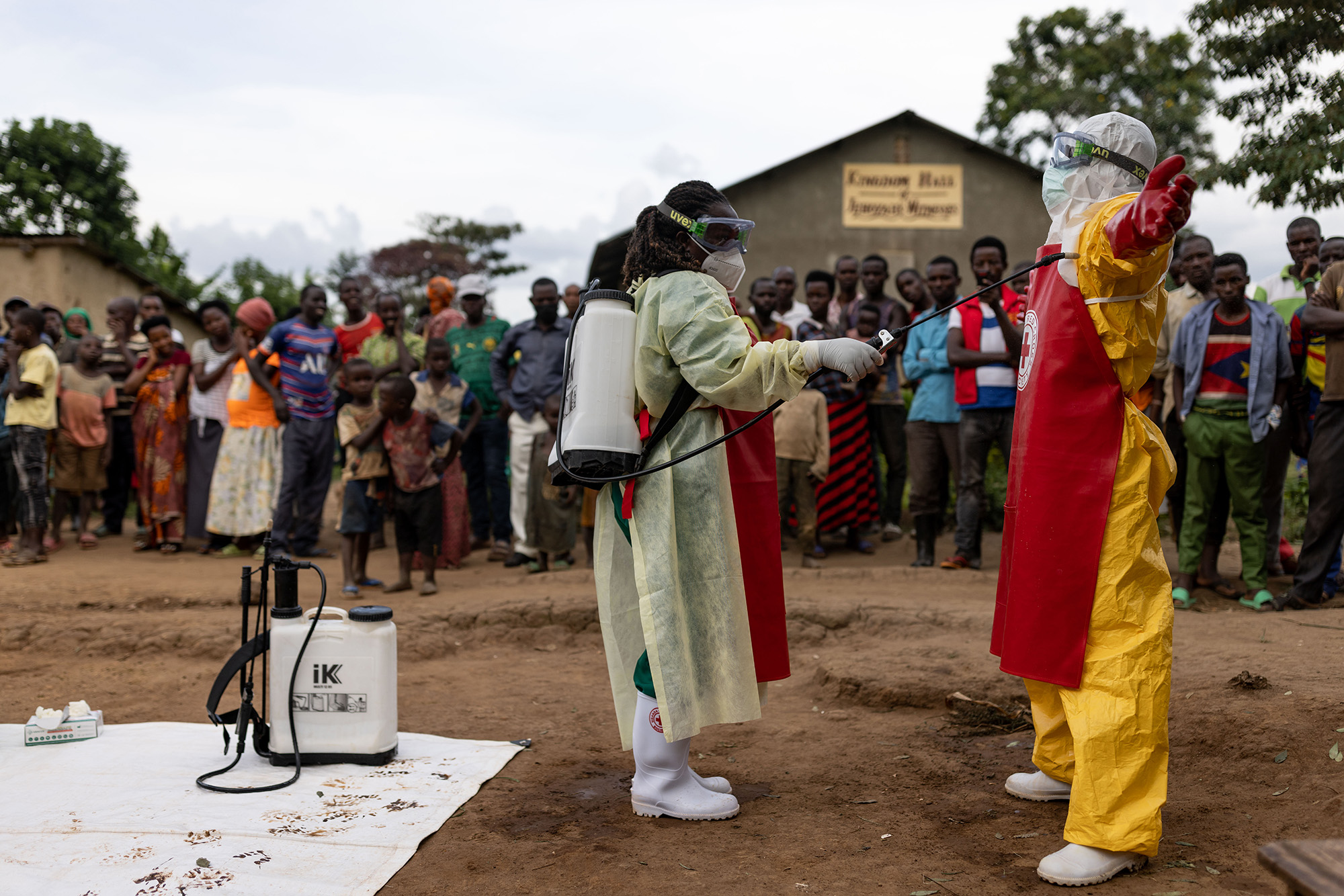 A health worker in PPE sprays another worker in PPE as villagers look on.