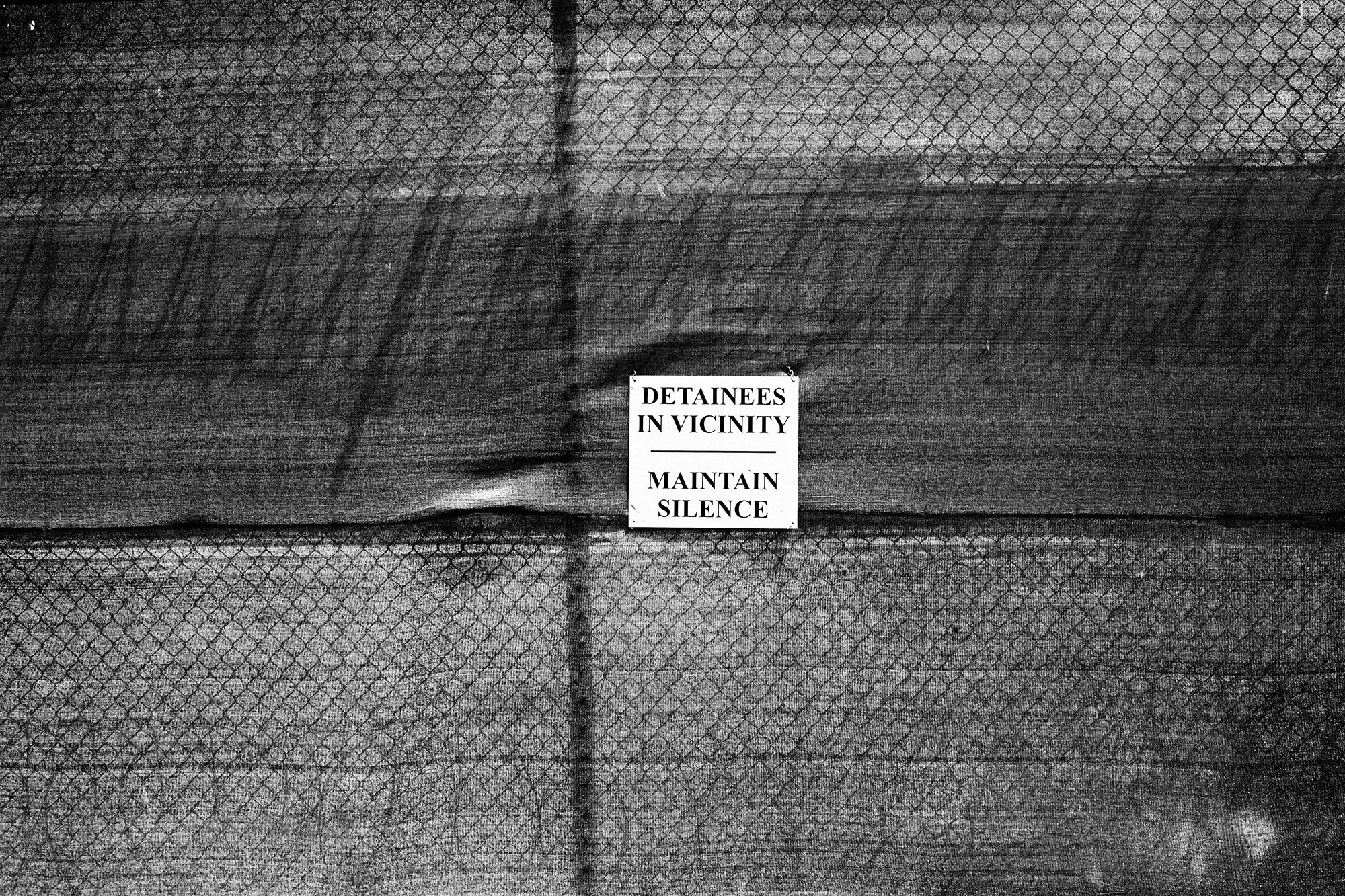 A sign hung on a chain-link fence reads “Detainees in vicinity. Maintain silence.”