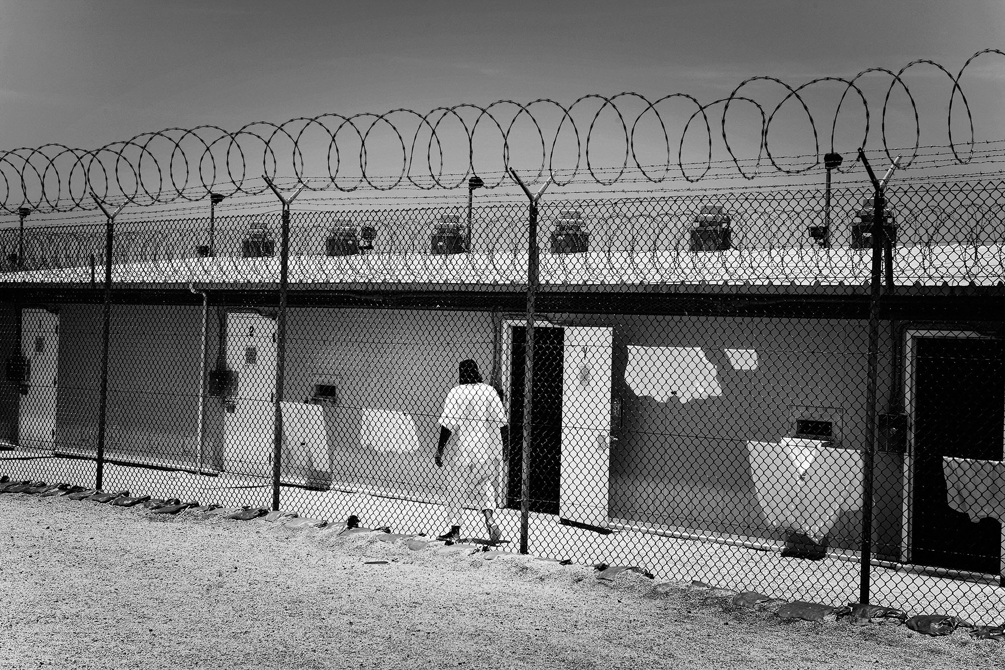 A detainee walks through an outdoor corridor bordered by a chain-link fence topped with razor wire.