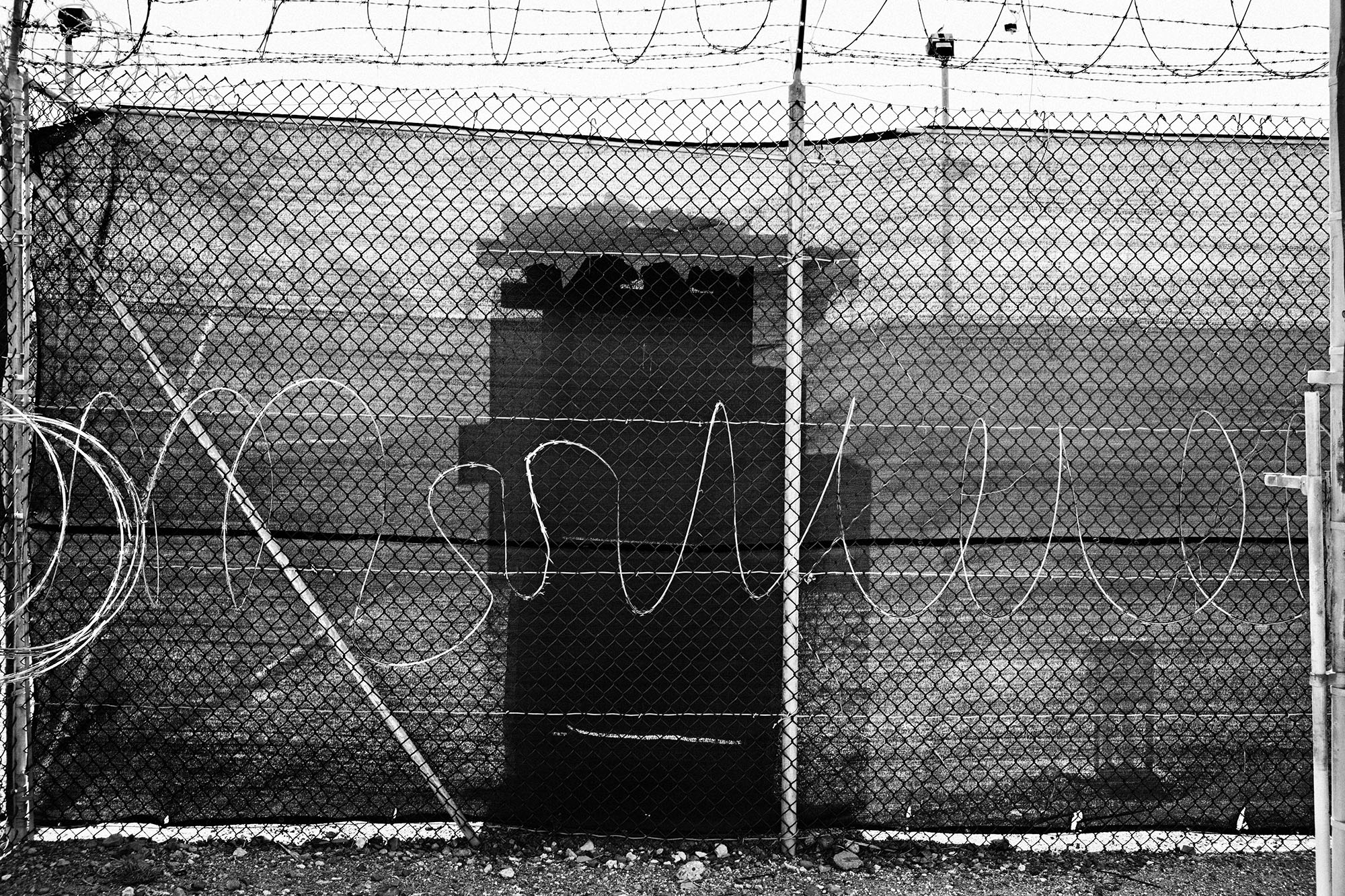 The silhouette of a guard tower is seen through a chain-link fence covered with tarp and topped with razor wire.