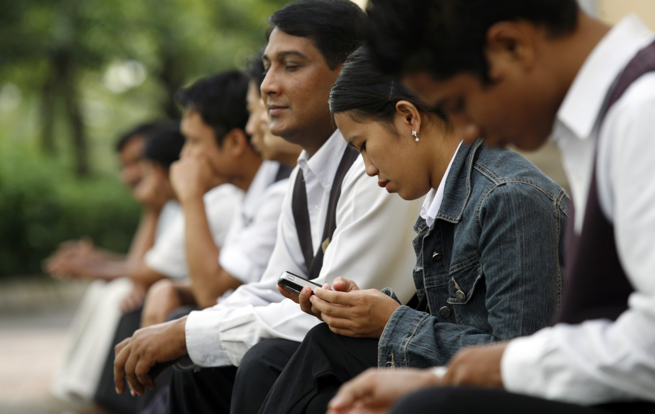 A group of workers from Bangladesh and Indonesia, most wearing white shirts and dark pants, sit in a line as they wait for public transportation.