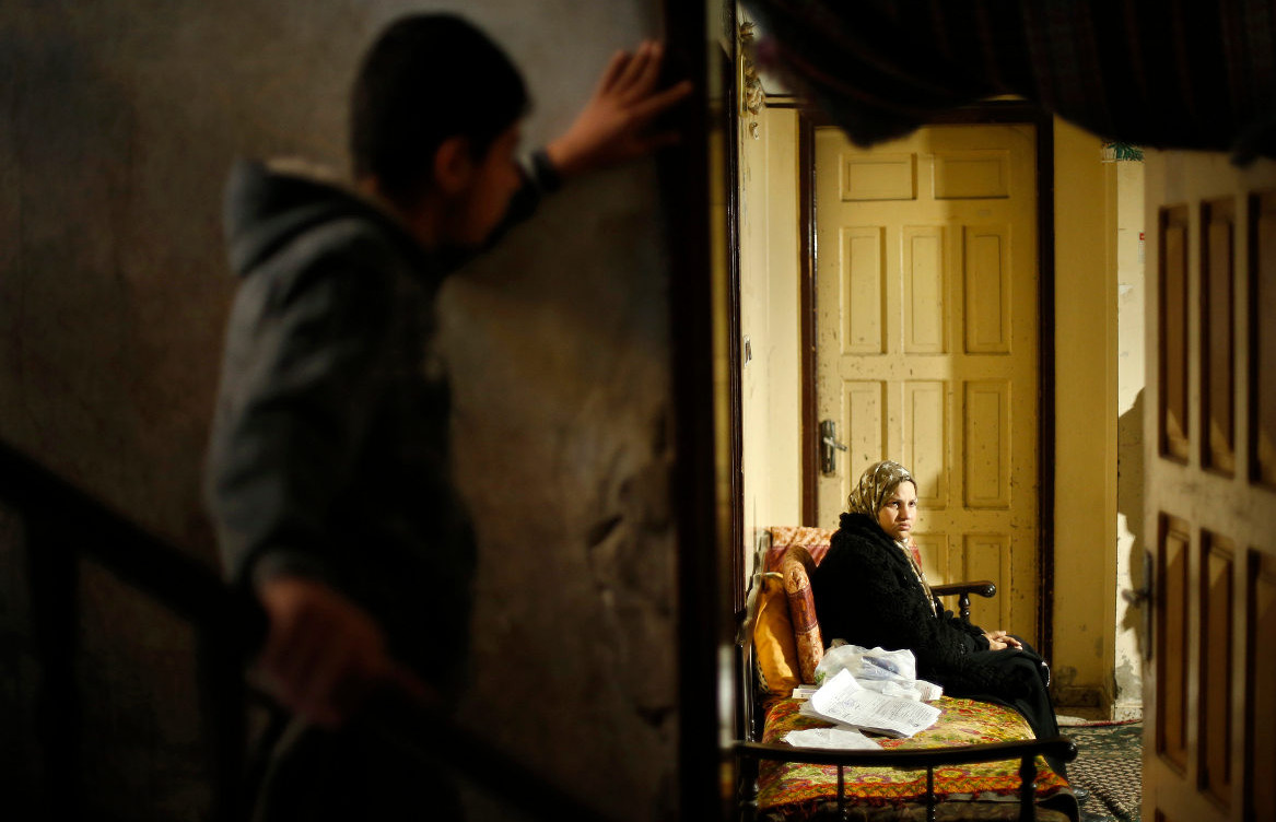 A boy looks on as a Palestinian woman suffering from cancer sits inside her home.