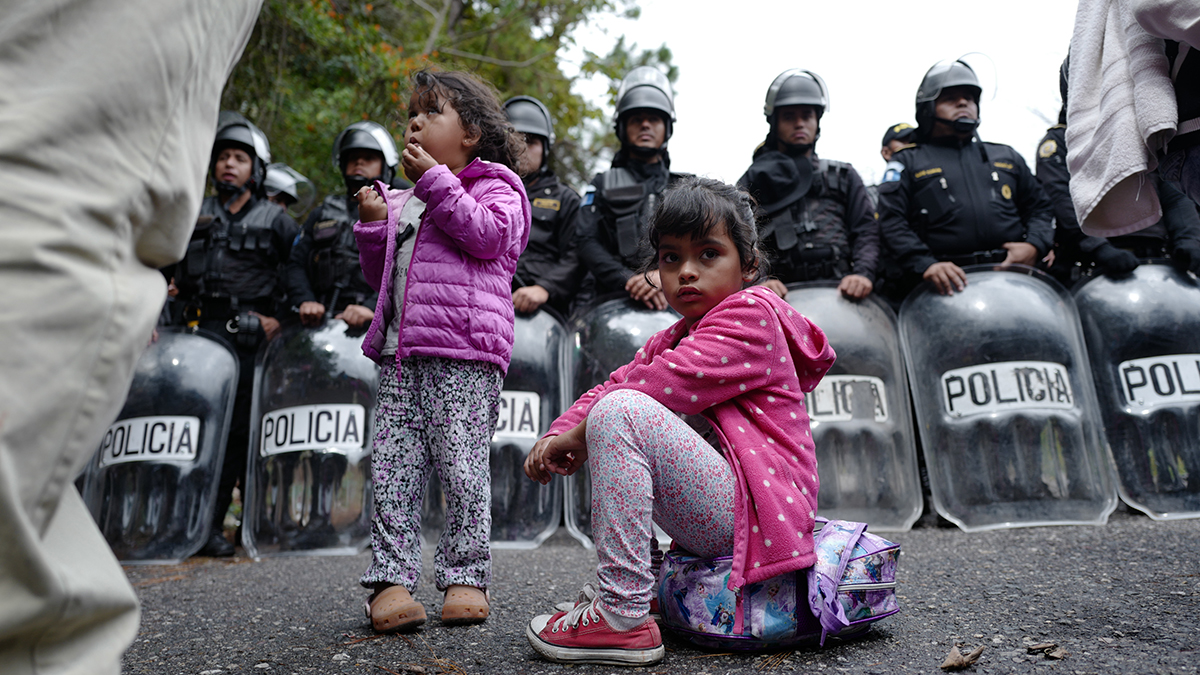 A migrant child sits on a backpack in front of police officers at the Aguas Calientes border crossing between Honduras and Guatemala.