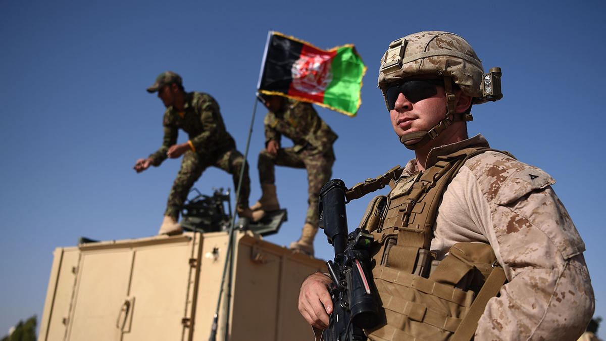 A U.S. Marine stands next to an armored vehicle as two Afghan troops raise a flag on top of it.