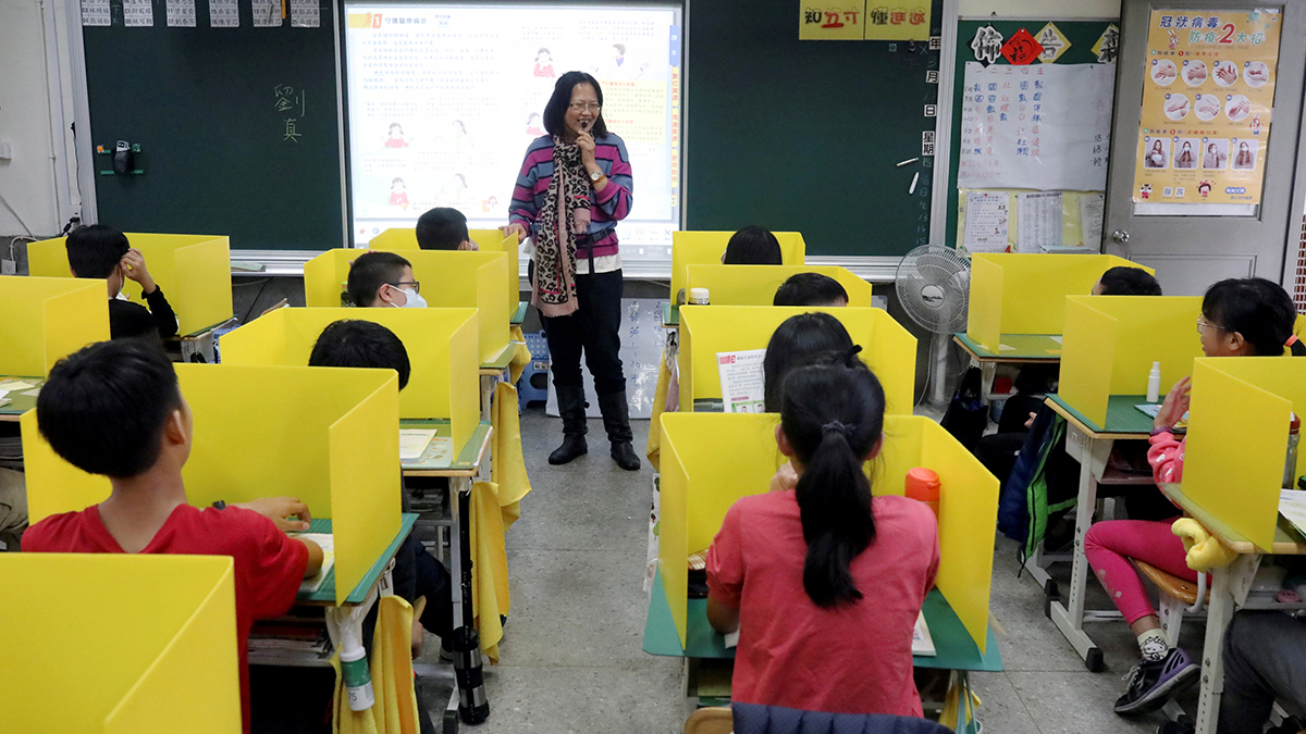 A teacher stands before several students in her classroom. Each of the students have dividers on their desks to separate them from other students. 