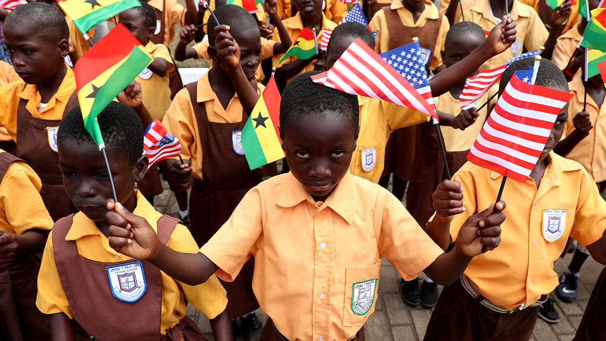 A crowd of Ghanaian children in brown and yellow school uniforms wave American and Ghanaian flags