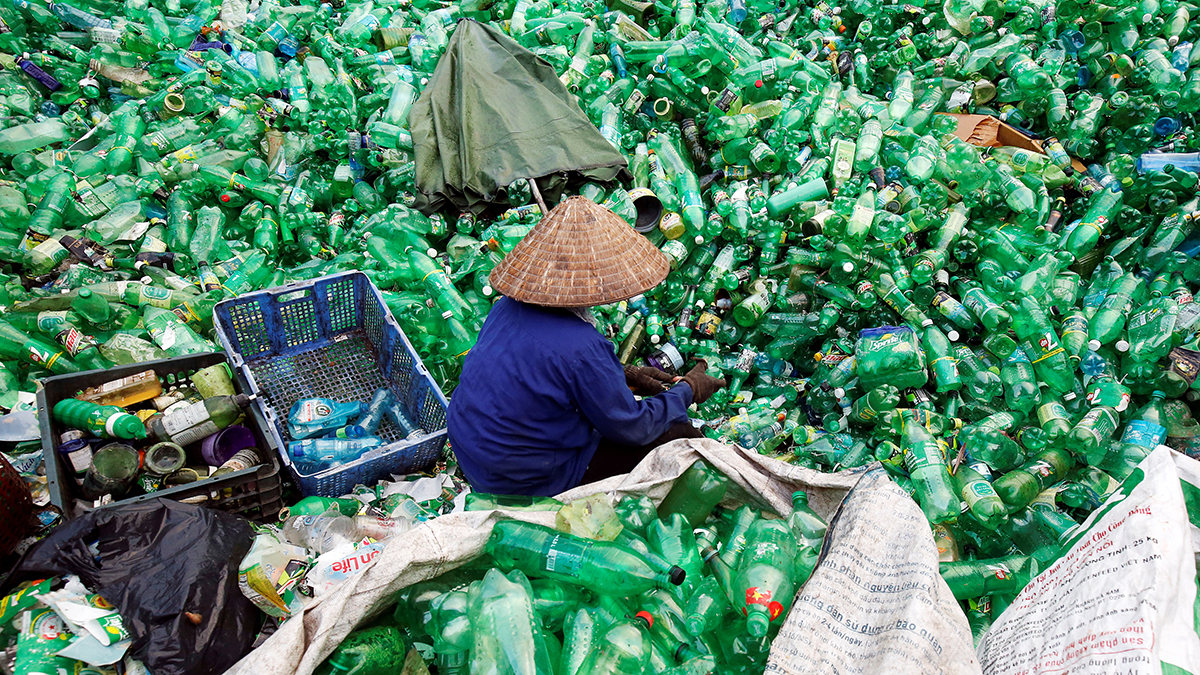 A woman in blue wearing a straw hat sitting amongst a sea of green soft drink plastic bottles. 