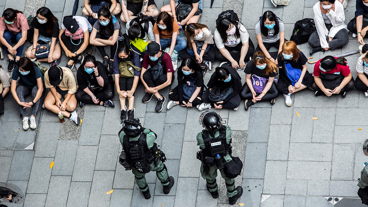 Two riot police officers stand by a group of detained protesters who sit on the ground.