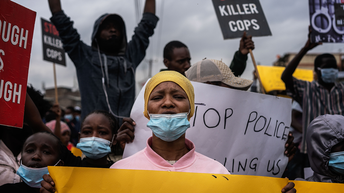 A woman looks anguished, wears a mask, and holds a sign amid other protesters holding signs that say 
