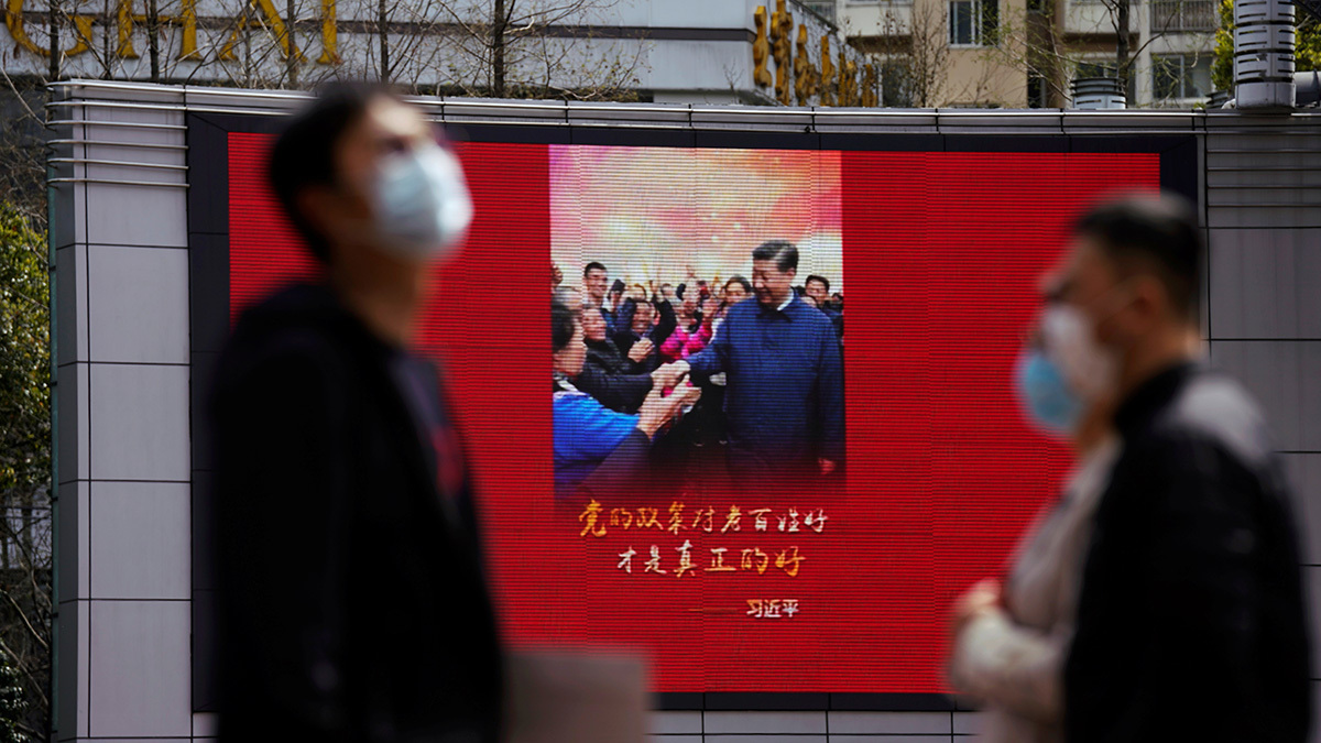 People wearing masks walk by a display showing an image of Xi Jinping smiling.