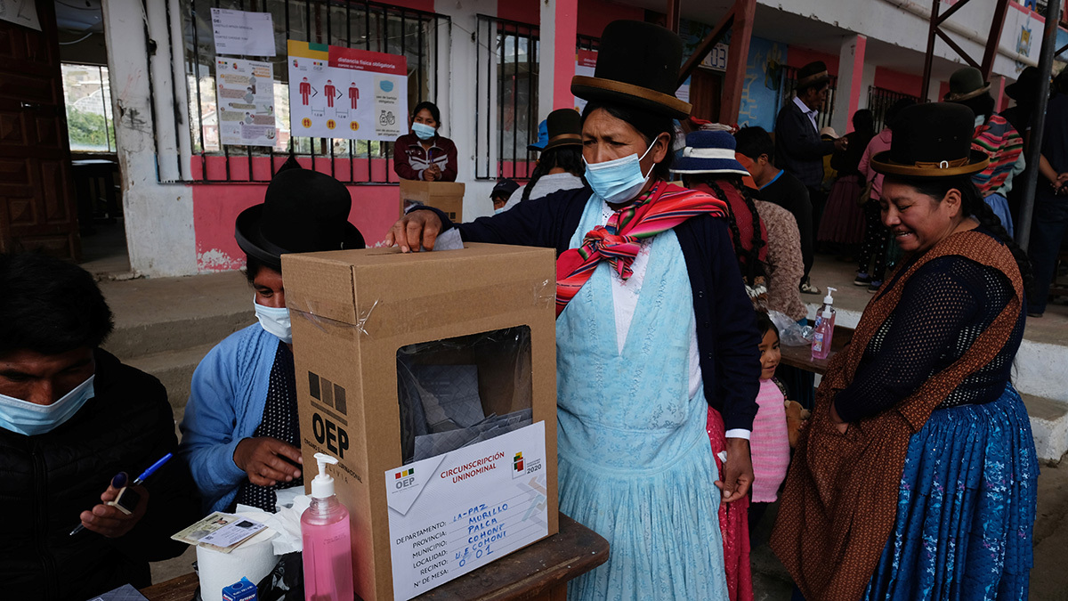 Women in skirts, aprons, and hats gather near a cardboard ballot box.  On of them, wearing a mask, is placing her ballot inside.