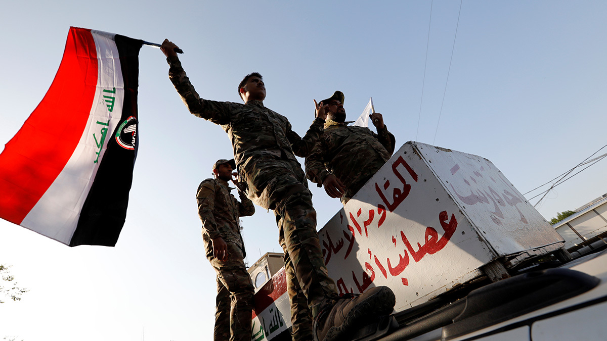 A member of Iraq's Popular Mobilization Forces waves a flag while standing on a vehicle with two other fighters