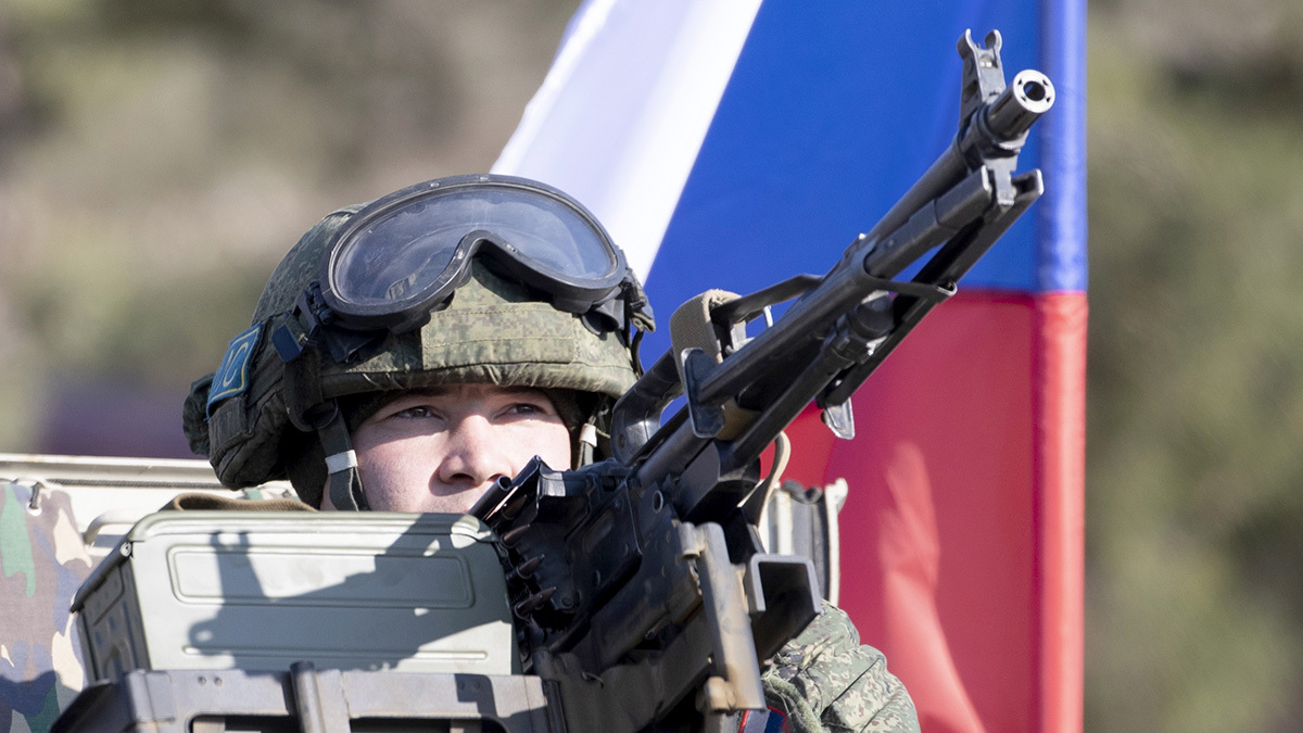 A Russian peacekeeper in Nagorno-Karabakh sits in a military vehicle and holds a gun. A Russian flag is in the background.