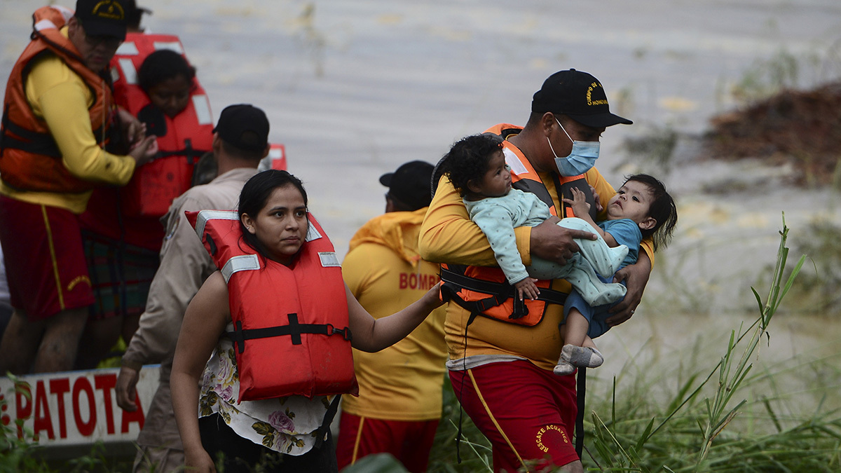 People, including a man cradling two babies, wearing orange life vests.  Water appears behind them.