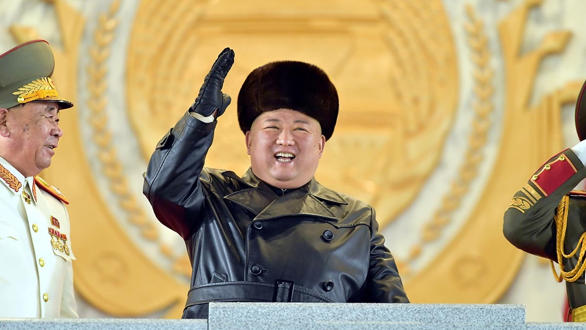 Kim Jong-un wears a leather jacket and fur hat. He waves his hand and smiles.