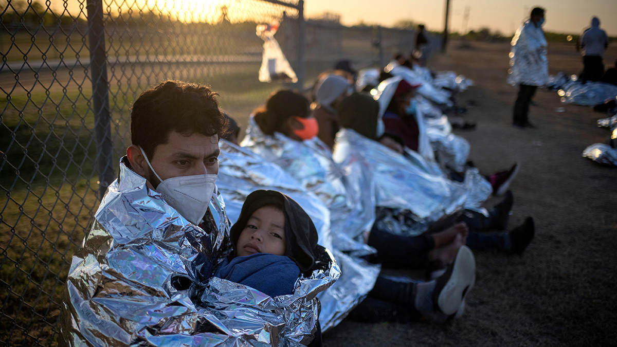 A man wearing a mask and wrapped in a shiny silver material holds a young child while sitting along a fence. Other people are in the background. It is sunrise.
