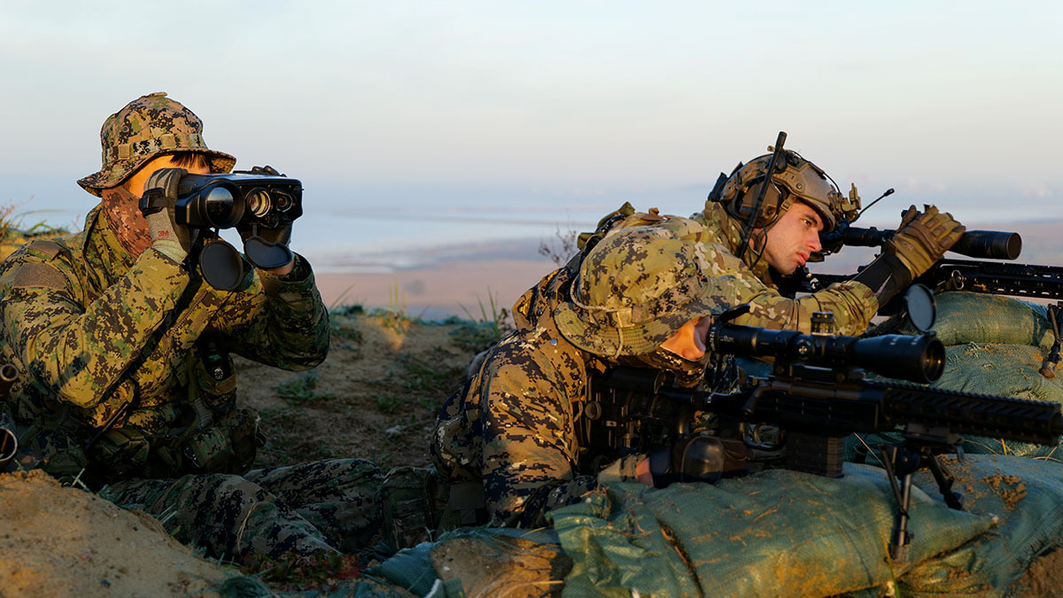Two people in military gear sit next to each other during a military exercise.