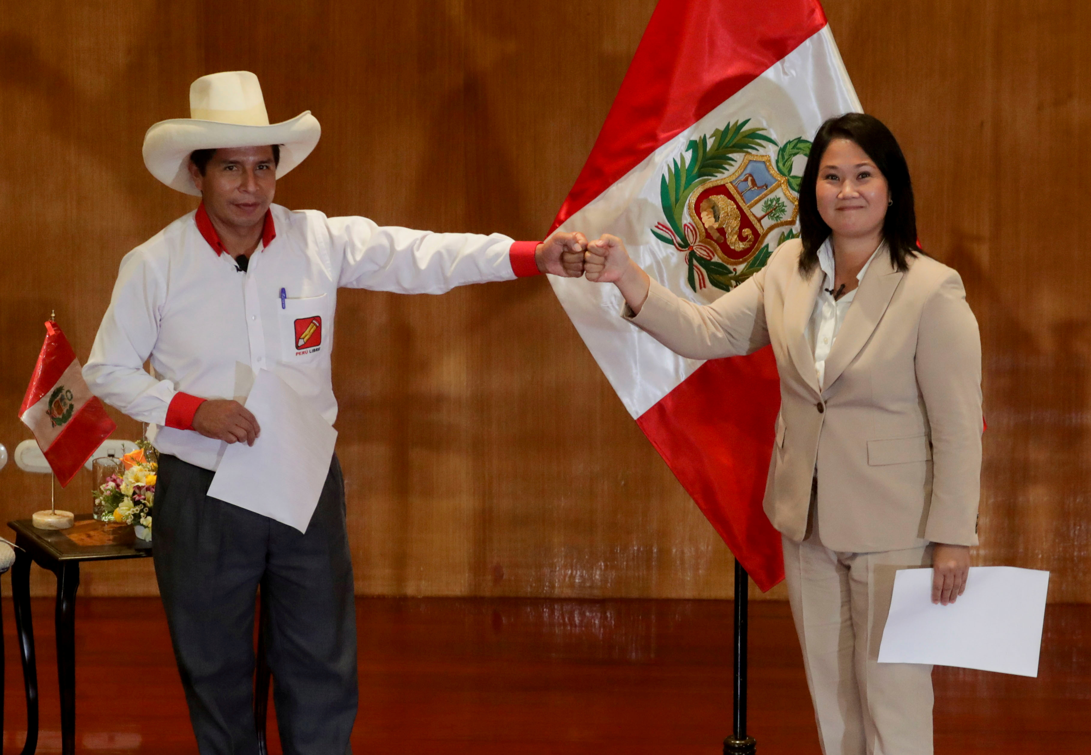 Pedro Castillo, dressed in a white shirt and wide-brimmed hat, fist bumps Keiko Fujimori, who is wearing a tan suit, in front of the Peruvian flag.