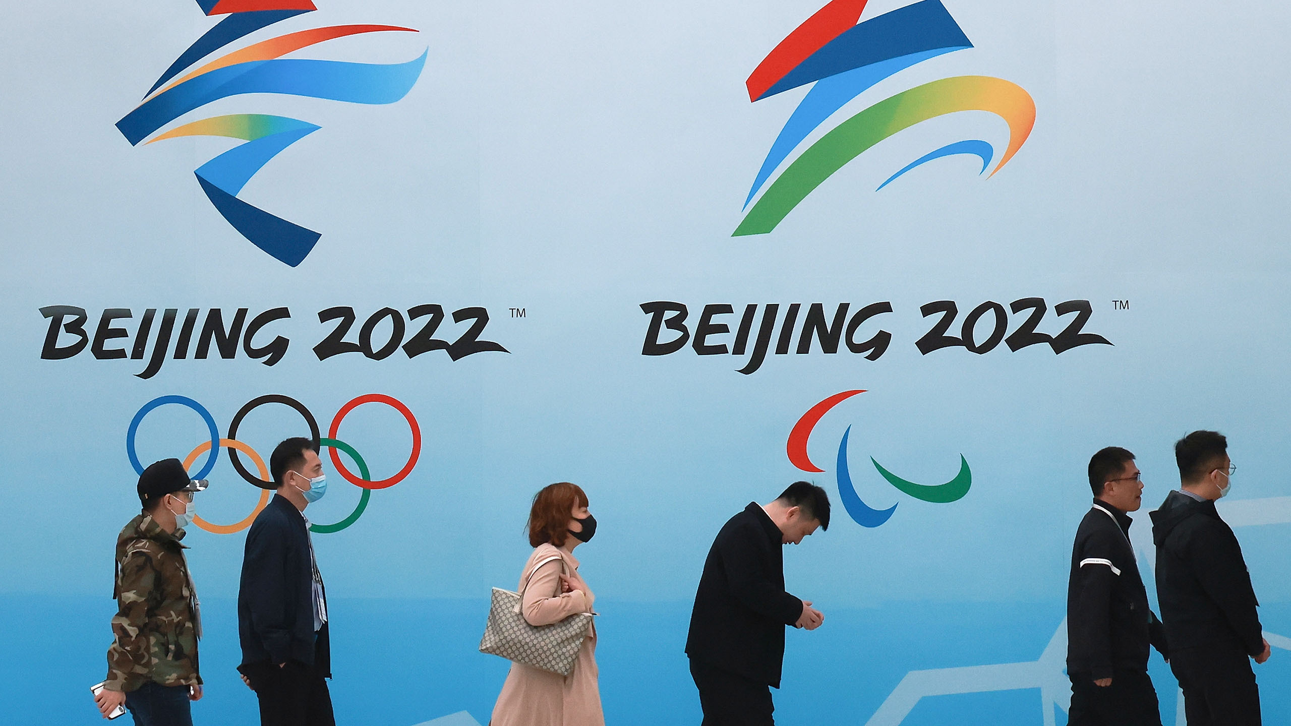 People wear protective masks as they walk in front of logos for the 2022 Beijing Winter Olympics.