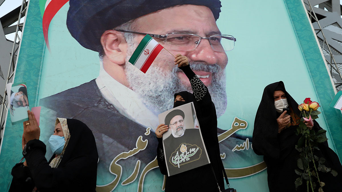 Three women in front of a mural of President-Elect Ebrahim Raisi. They hold flowers, Iranian flags, and his portrait.