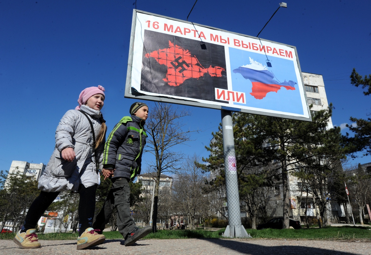 Children walk past a billboard showing one Crimea with a swastika and barbed wire and one with the colors of the Russian flag