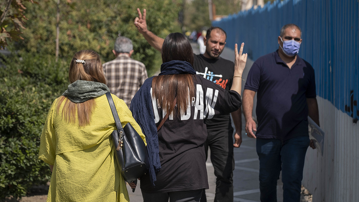Two women without headscarves walk along a Tehran street. A man and one of the women flash victory signs at each other.