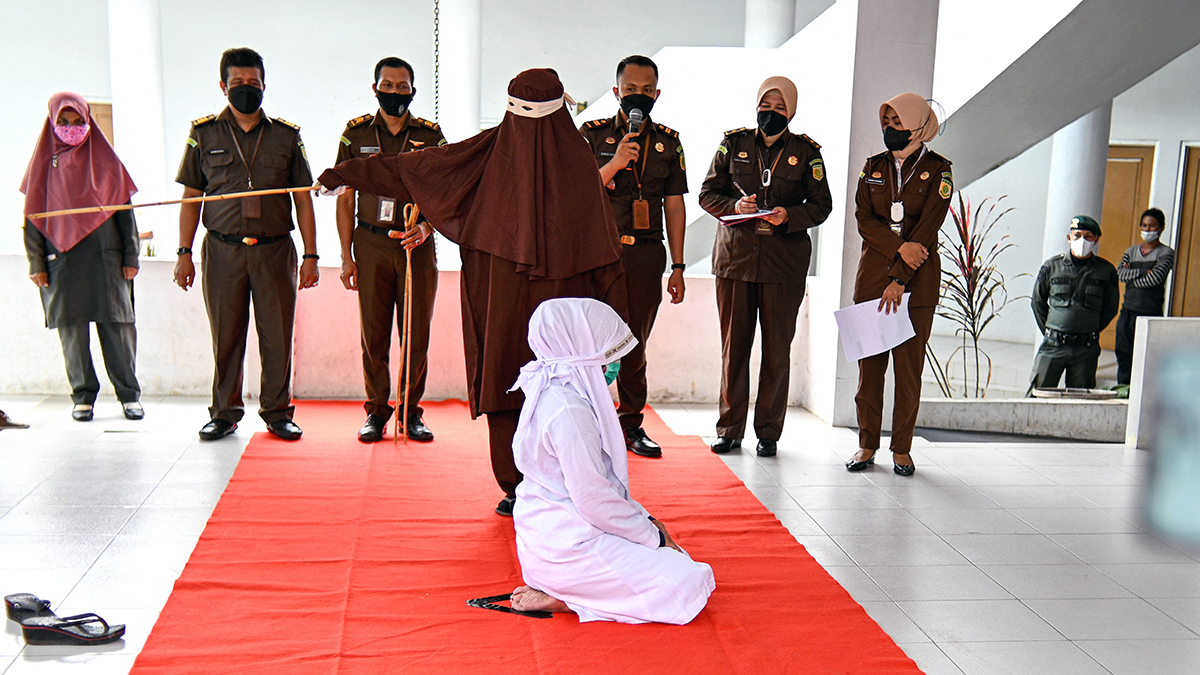 A woman in white kneels as a woman sharia officer swings a cane to hit her. Officers and a spectator watch in the background.