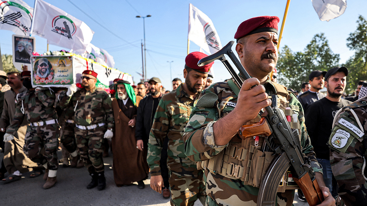 A group of armed militants march in uniform during a funeral procession, carrying a coffin of a fallen troop and raising flags.