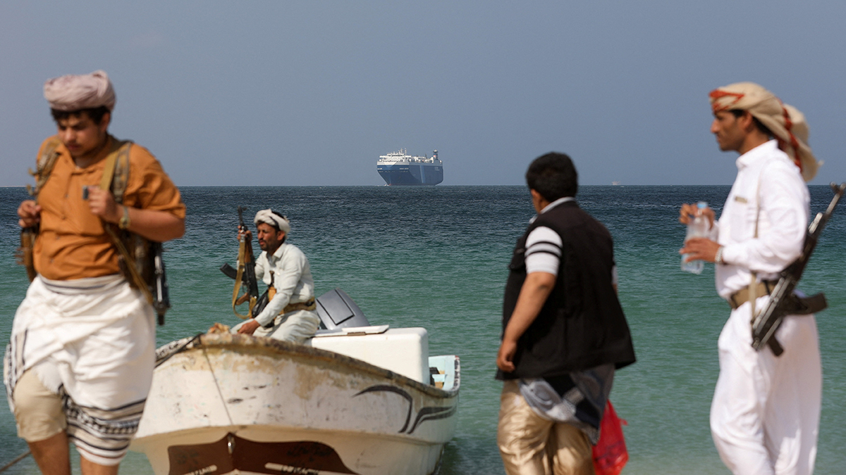 Armed men pull a small boat ashore, as a commercial vessel looms in the background.