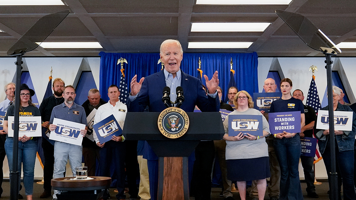 President Joe Biden stands at a podium in front of members of the steelworkers union holding placards.