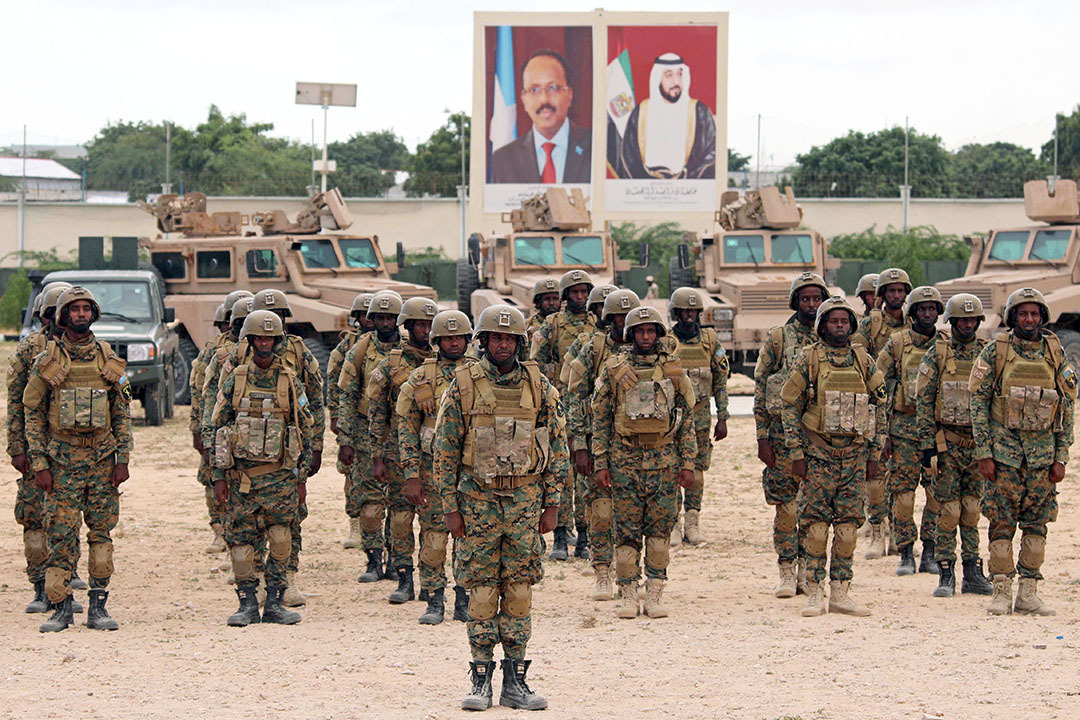 Soldiers stand in rows on a gravel lot, with large portraits of leaders behind them.
