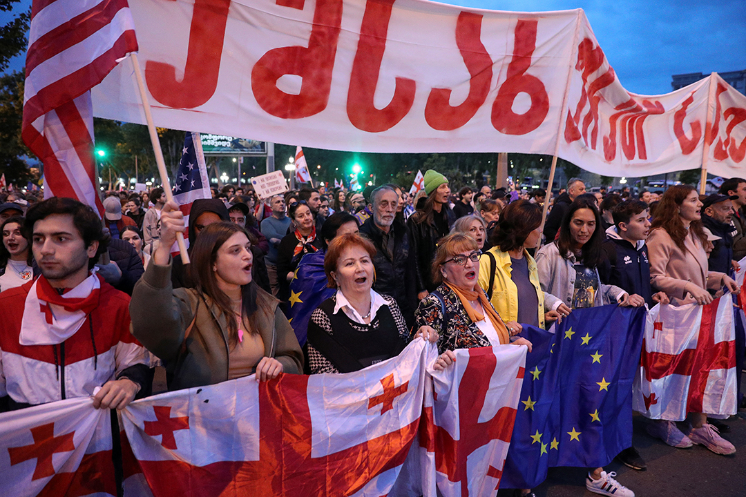 A crowd of protestors holding Georgian and EU flags raise a banner in Georgian script.
