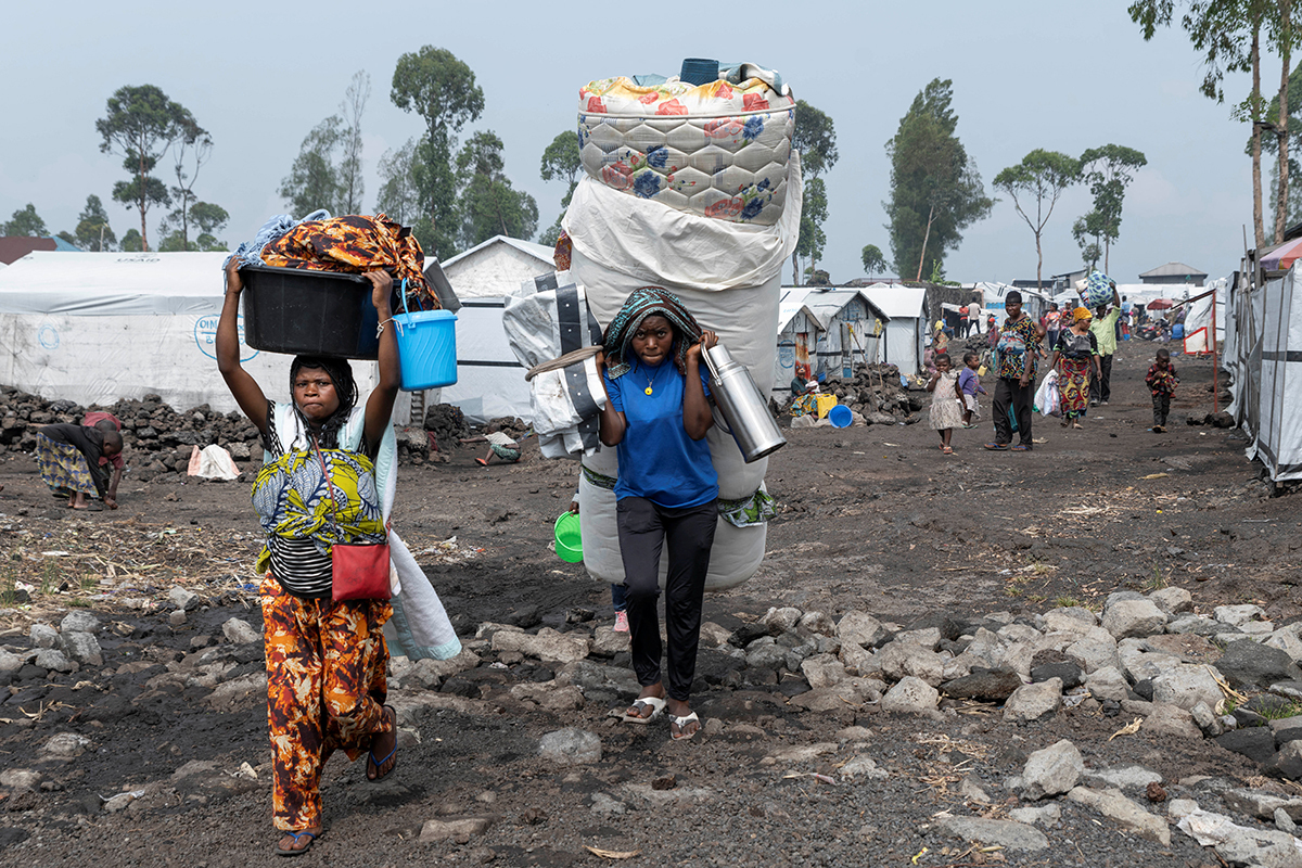 Refugees carry belongings, including a mattress and other supplies across a refugee camp.