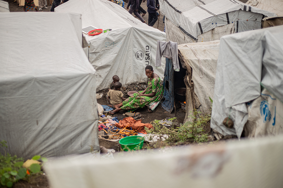 A woman sits on the ground surrounded by white humanitarian relief tents. 