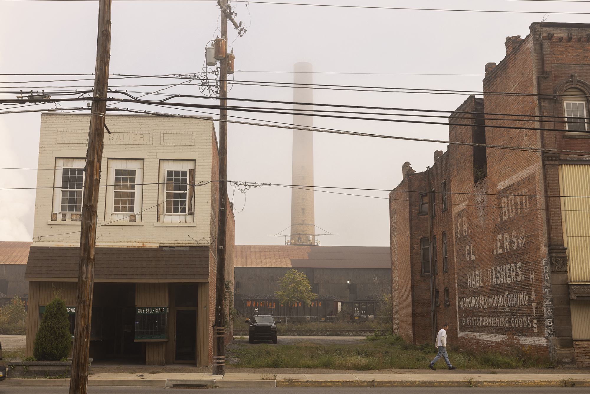 A man walking in front of an empty lot between two buildings. A smokestack is visible in the background.