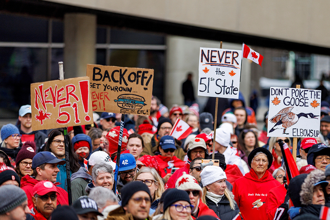 A group of people dressed in the colors of Canada—typically red and white—are gathered. They are holding up signs that in protest of Trump's comments about Canada as the 51st state.