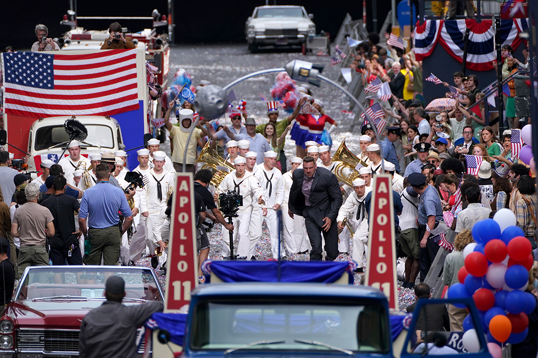 Actors in sailor costumes rush toward an arch on set surrounded by props including American flags, balloons, and 