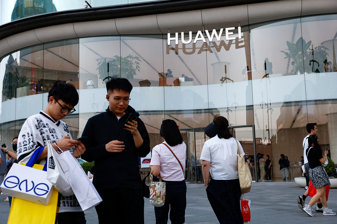 Two men look at their phones outside a glass building with the name Huawei affixed to it. 