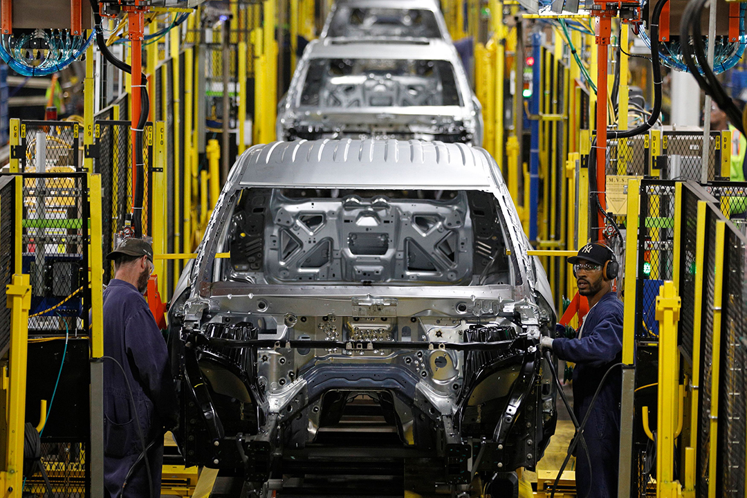 Workers in blue jumpsuits assemble cars on an assembly line 