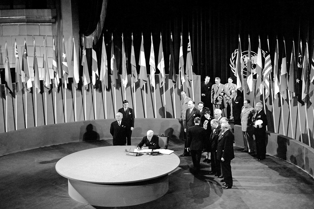 A black-and-white image. A man in a suit sits at a table surrounded by flags. He is signing a document. Men in suits and military garb watch as the ceremony proceeds.